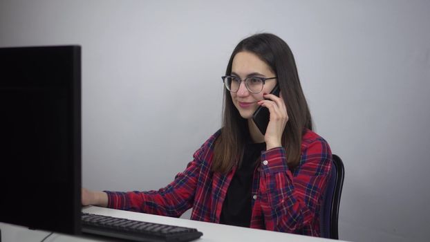 IT specialist sits at the workplace in the office and speaks on the phone. A young woman in glasses and a red shirt is working. 4k