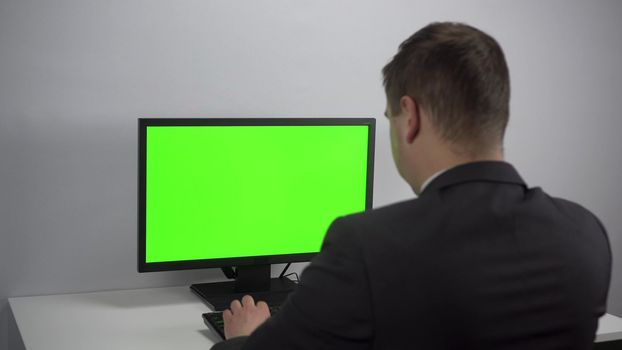 A young businessman is sitting in front of a computer with a green screen. A man in a suit works for a PC. Screen with chroma key. 4k