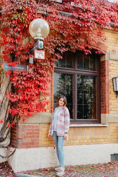 a beautiful girl stands against the background of the window of an old European house, entwined with a floating red color in autumn. Tourism & Travel Concept. Nice portrait of a young woman, in boho style outdoors in fall autumn day. Caucasian female girl 20 30 years old