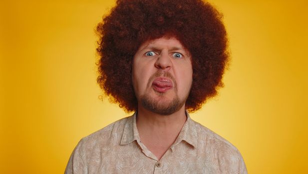 Funny joyful sincere man with lush Afro hairstyle coiffure making playful silly facial expressions and grimacing fooling around showing tongue. Young handsome hipster guy isolated on yellow background