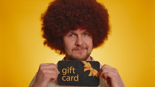 Young man with lush afro hairstyle showing pointing on gift discount certificate voucher coupon for store holidays sale. Hipster guy with shopping Christmas surprise card isolated on yellow background