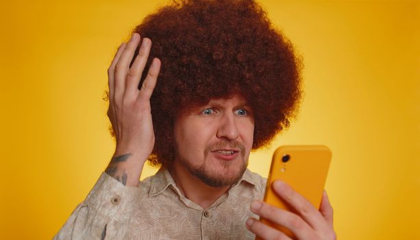 Bearded man with lush afro hairstyle coiffure use mobile cell phone typing browsing, loses becoming surprised sudden lottery results, bad fortune, loss, fail. Young guy isolated on yellow background