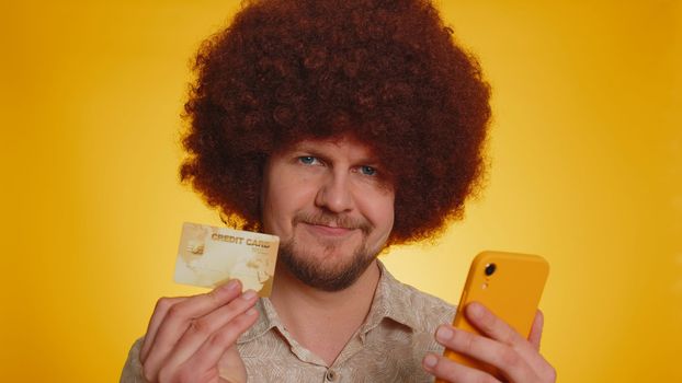 Sincere man with lush afro hairstyle customer using credit bank card and smartphone while transferring money purchases online shopping, payment. Finance and internet. Young guy on yellow background