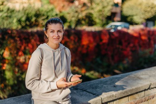 Young fashionable teenage girl with smartphone in park in autumn sitting at smiling. Trendy young woman in fall in park texting. Retouched, vibrant colors. Beautiful blonde teenage girl wearing casual modern autumn outfit sitting in park in autumn. Retouched, vibrant colors, brownish tones.