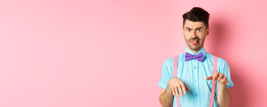 Funny guy looking at something disgusting with aversion and cringe, show tongue and shaking hands in disapproval, standing on pink background.