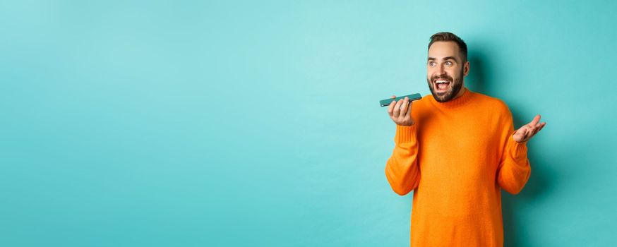 Happy man talking on speakerphone, gesturing and recording voice message on mobile phone, standing in orange sweater over light blue background.
