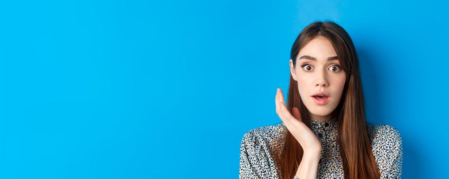 Candid girl with long hair and makeup, looking shocked and surprised, open mouth and stare speechless at camera, standing on blue background.