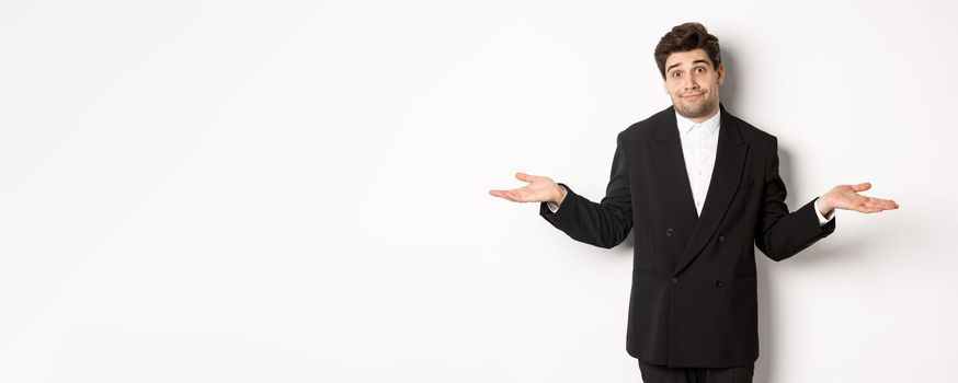Image of handsome confused man in black suit, shrugging and looking clueless, dont know anything, standing over white background.