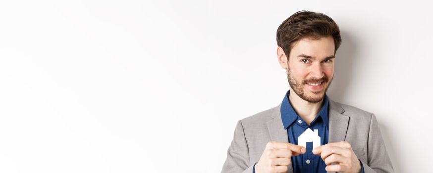 Real estate and insurance concept. Smiling male realtor showing small paper house cutout and looking at camera, selling property, white background.