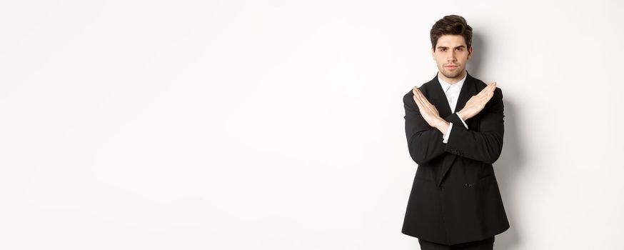 Portrait of serious and concerned man in black suit, showing stop gesture and frowning, making cross to prohibit or decline something, standing over white background.