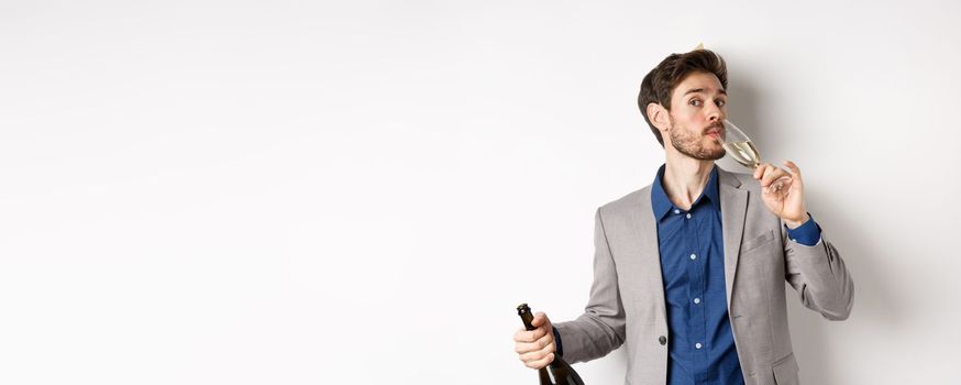 Celebration and holidays concept. Handsome bearded man in suit and birthday hat holding bottle, drinking glass of champagne, standing on white background.