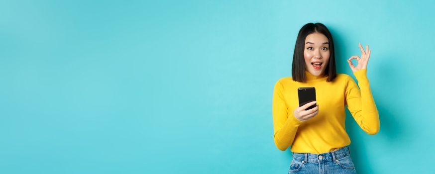 E-commerce and online shopping concept. Portrait of asian woman showing OK sign and using mobile phone, recommend application, standing over blue background.