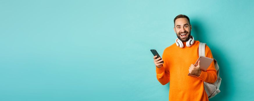 Handsome man student with headphones and backpack, holding digital tablet and smartphone, looking at camera, standing against turquoise background.