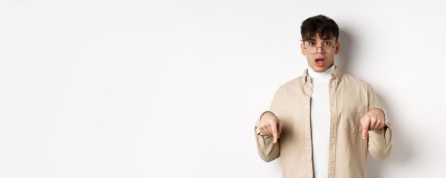 Shocked attractive young man showing something awesome, drop jaw and gasping in awe, pointing fingers down, standing on white background.