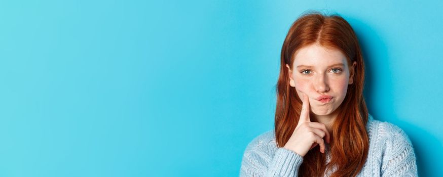 Puzzled redhead girl looking suspicious at camera, thinking or solving problem, standing against blue background.