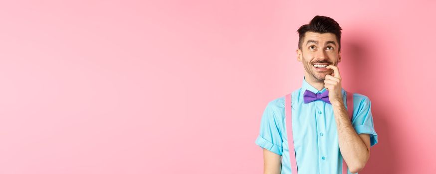 Image of smiling man making choice, looking dreamy and happy up, biting fingernail with tempted expression, wanting something, standing over pink background.