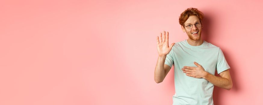 Friendly redhead man being honest, holding hand on heart and arm raised high to swear or make promise, smiling at camera, telling truth over pink background.
