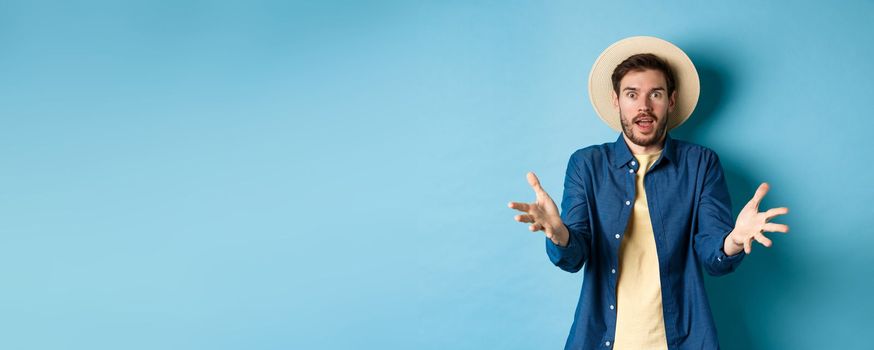 Surprised young man in summer hat, raising hands up staring with disbelief and amazement at camera, impressed with big event, standing on blue background.