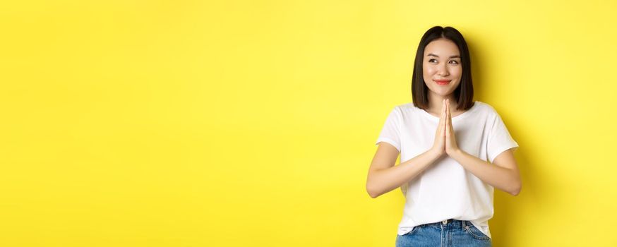 Pretty asian woman holding hands in namaste, pray gesture, looking left and smiling, say thank you, express gratitude, standing over yellow background.