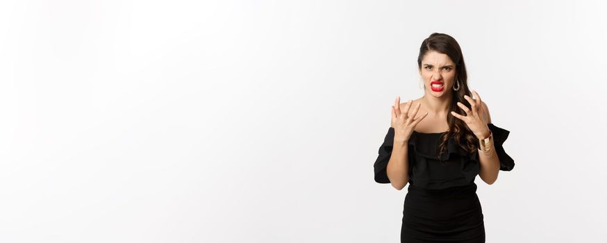 Fashion and beauty. Angry woman in black dress, shouting mad and shaking hands, grimacing outraged at camera, standing over white background.