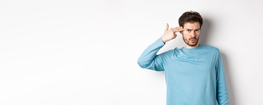 Annoyed young man frowning, showing shooting gun near head gesture, blow brain out sign, standing over white background.