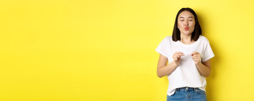 Young asian woman in casual white t-shirt showing plastic credit card and pucker lips silly, going on shopping, yellow background.