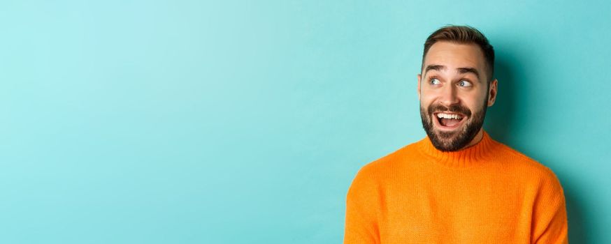 Close-up of handsome caucasian man smiling, looking left with surprised face, staring at logo, wearing orange sweater, standing against turquoise background.