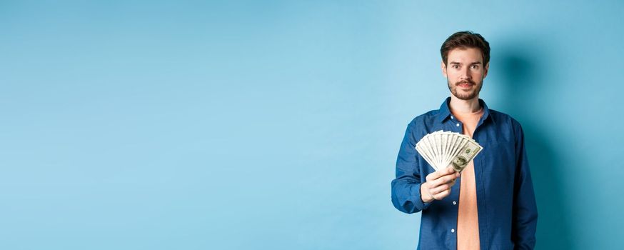 Happy modern guy holding money and smiling, standing on blue background.