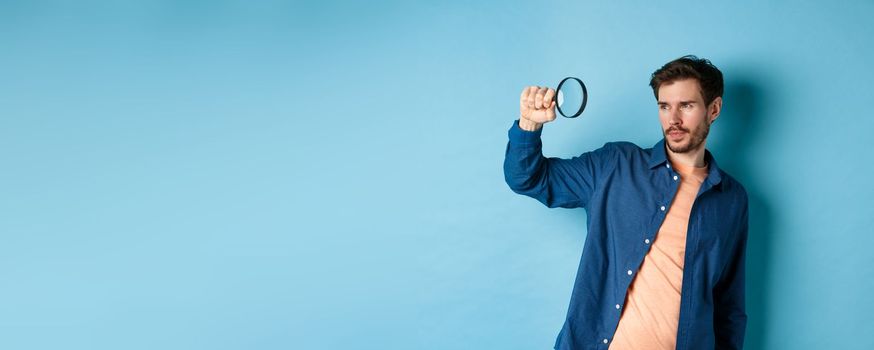 Image of young man searching for something, looking at empty space with magnifying glass, standing on blue background.