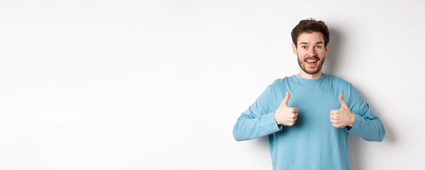 Excited smiling man showing thumbs up to praise excellent product, looking amazed and happy at camera, recommending good deal, standing on white background.