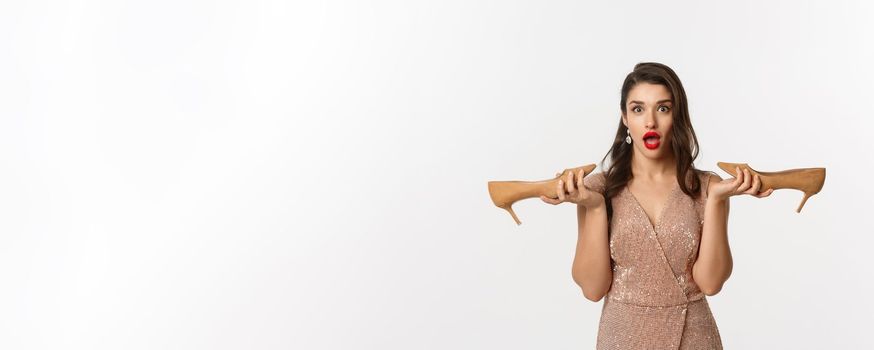 Party and celebration concept. Attractive woman in elegant dress holding pair of heels and looking surprised, dressing up for Christmas, white background.