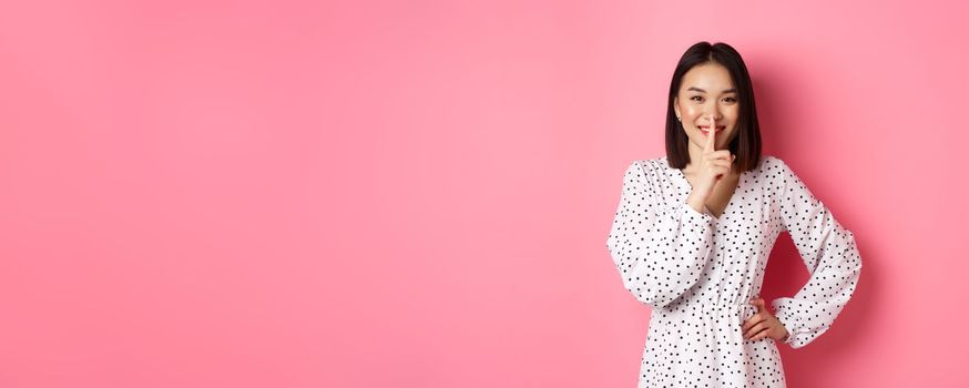 Beautiful Korean woman in trendy dress asking to keep secret, hushing with soft smile, planning a surprise, standing over pink background.