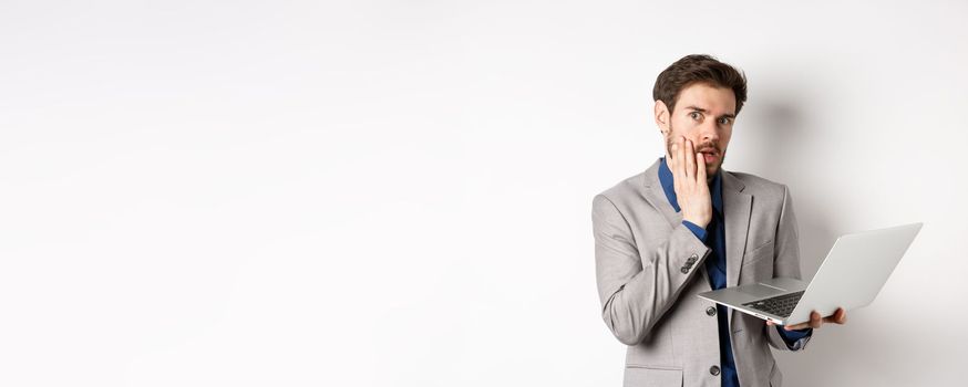 Shocked and troubled businessman have trouble on laptop, looking worried at camera, made mistake at work, standing on white background in suit.