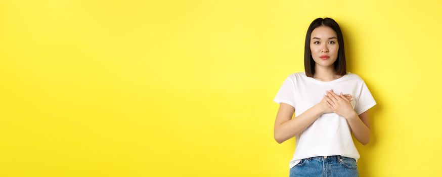 Beauty and fashion concept. Beautiful asian woman holding hands on heart and looking thoughtful at camera, keeping memories in soul, standing over yellow background.