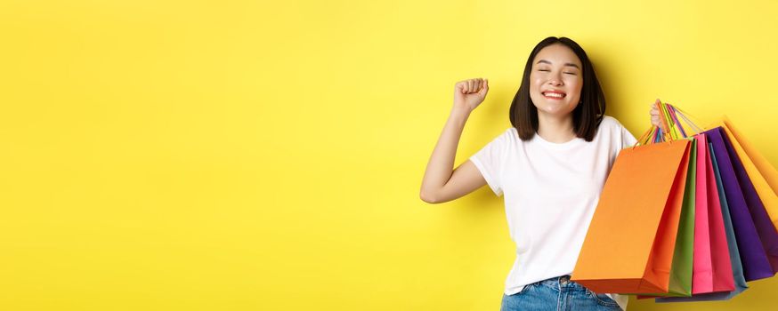 Happy asian woman feeling satisfied after shopping on sale, holding paper bags and stretching with pleased smile, standing over yellow background.