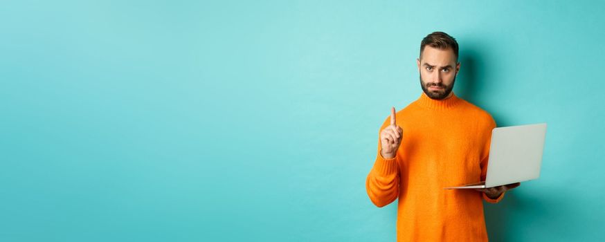 Image of adult man teaching online, showing finger while using laptop, explaining rules, standing over light blue background.