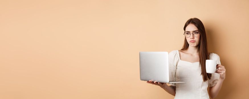 Tired working girl looking at laptop bored, drinking coffee while using computer for work, standing on beige background.