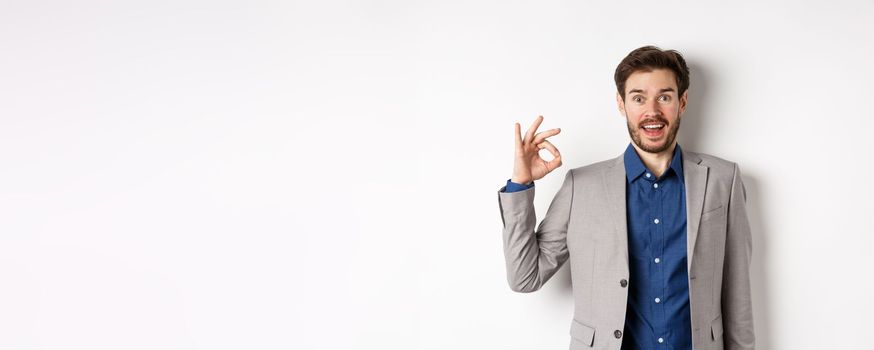 Excited bearded guy in business suit gasping amazed, looking with admiration and showing okay sign, say yes, approve and like good product, standing on white background.