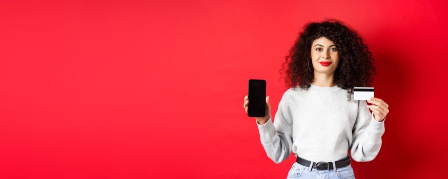 Young modern woman with curly hair showing plastic credit card and mobile phone screen, demonstrating online shopping app, standing on red background.