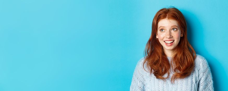 Close-up of beautiful redhead girl in sweater, looking at upper left corner promo logo and smiling, standing over blue background.