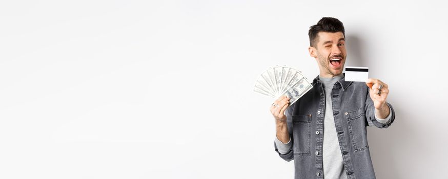 Image of handsome guy holding dollar bills but suggesting use plastic credit card, smiling friendly at camera, standing on white background.