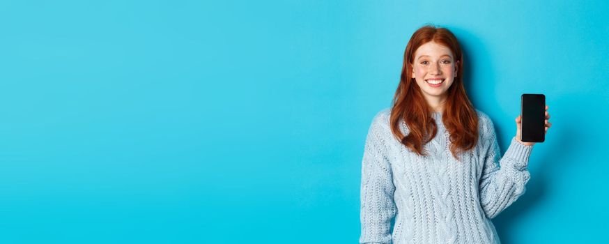 Smiling female model with red hair showing smartphone screen, holding phone and demonstrating application, standing over blue background.