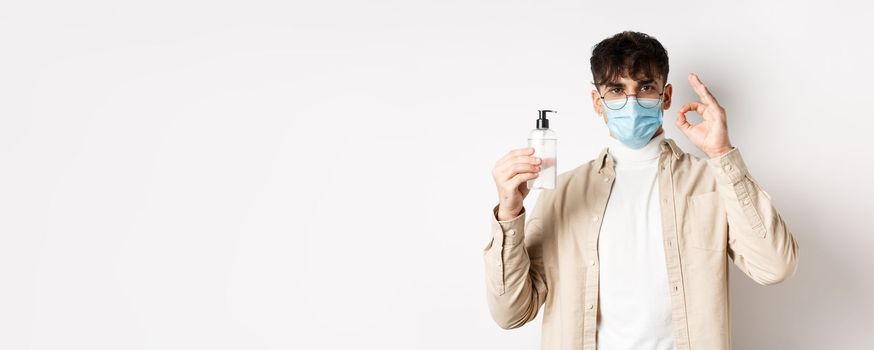 Health, covid and quarantine concept. Portrait of natural guy in glasses and face mask showing bottle of good hand sanitizer, make okay sign, recommending product, white background.