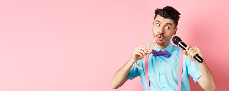 Party and festive events concept. Funny guy singing karaoke, performing song with microphone and drinking champagne from glass, standing on pink background.