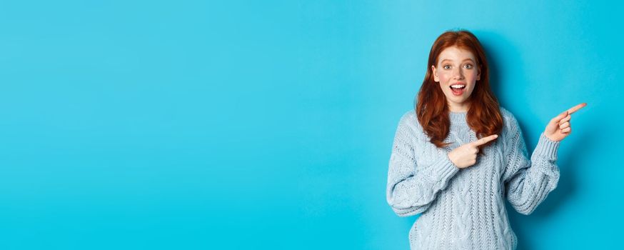 Winter holidays and people concept. Curious teenage girl in sweater, pointing fingers right and staring at camera amazed, showing promo offer, standing over blue background.