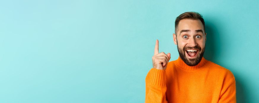 Close-up of handsome man having an idea, raising finger up and smiling excited, standing in orange sweater.