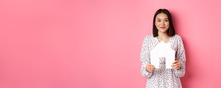 Real estate. Adult asian woman searching for home, holding house model and smiling, promo of broker company, standing over pink background.