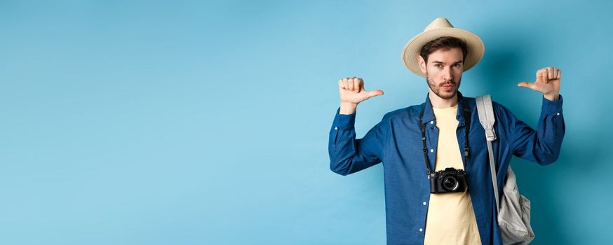 Confident handsome guy in summer hat, pointing at himself with bragging look, going to travel on holidays, holding backpack and camera, blue background.