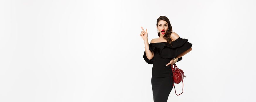 Beauty and fashion concept. Full length of excited young woman in glamour dress, red lips, having an idea, raising finger to suggest something, white background.