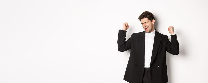 Image of successful and pleased handsome man in suit, rejoicing and making fist pump signs, dancing from happiness, standing over white background.